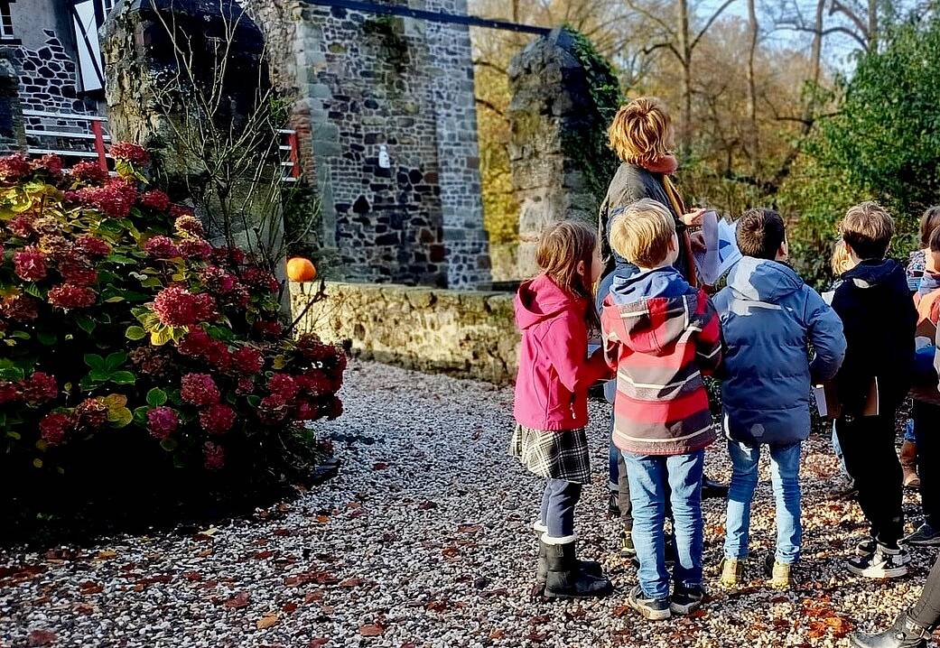 Das Bild zeigt eine Gruppe von Grundschulkindern und eine Frau vor altem Gemäuer, auf dem Boden Herbstlaub